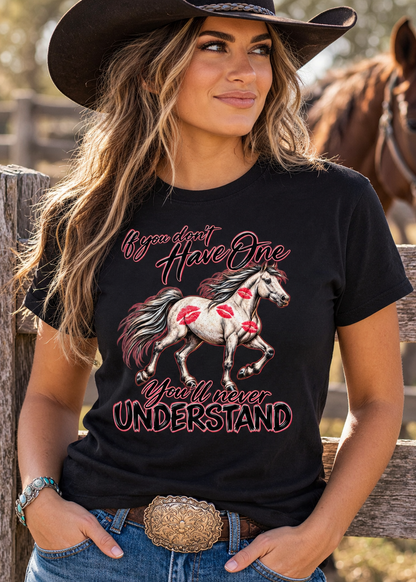 A woman wearing a black graphic t-shirt with a western design, featuring a white horse with red markings and the text 'If you don't Have One You will never UNDERSTAND'. She is also wearing a black cowboy hat and blue jeans, standing outdoors in front of a wooden fence with a horse in the background.
