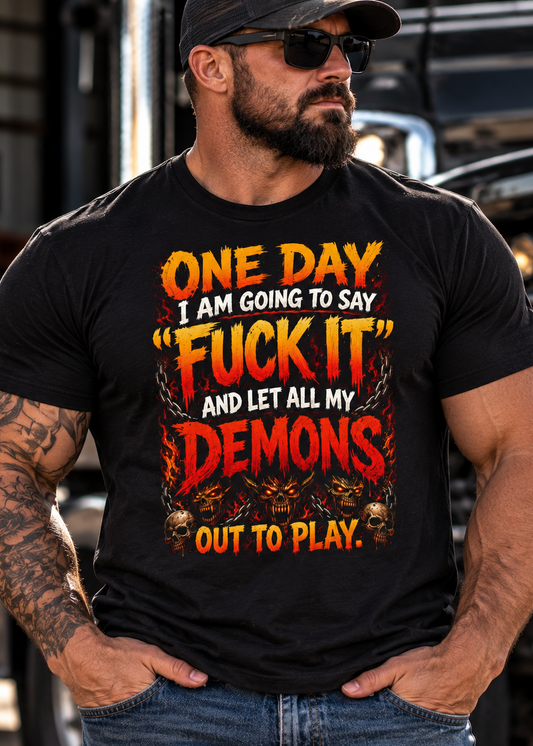 A muscular man wearing a black graphic t-shirt with a bold red and yellow text design that reads 'ONE DAY I AM GOING TO SAY "FUCK IT" AND LET ALL MY DEMONS OUT TO PLAY.' He is also wearing a black cap and sunglasses, standing in front of a blurred truck background.