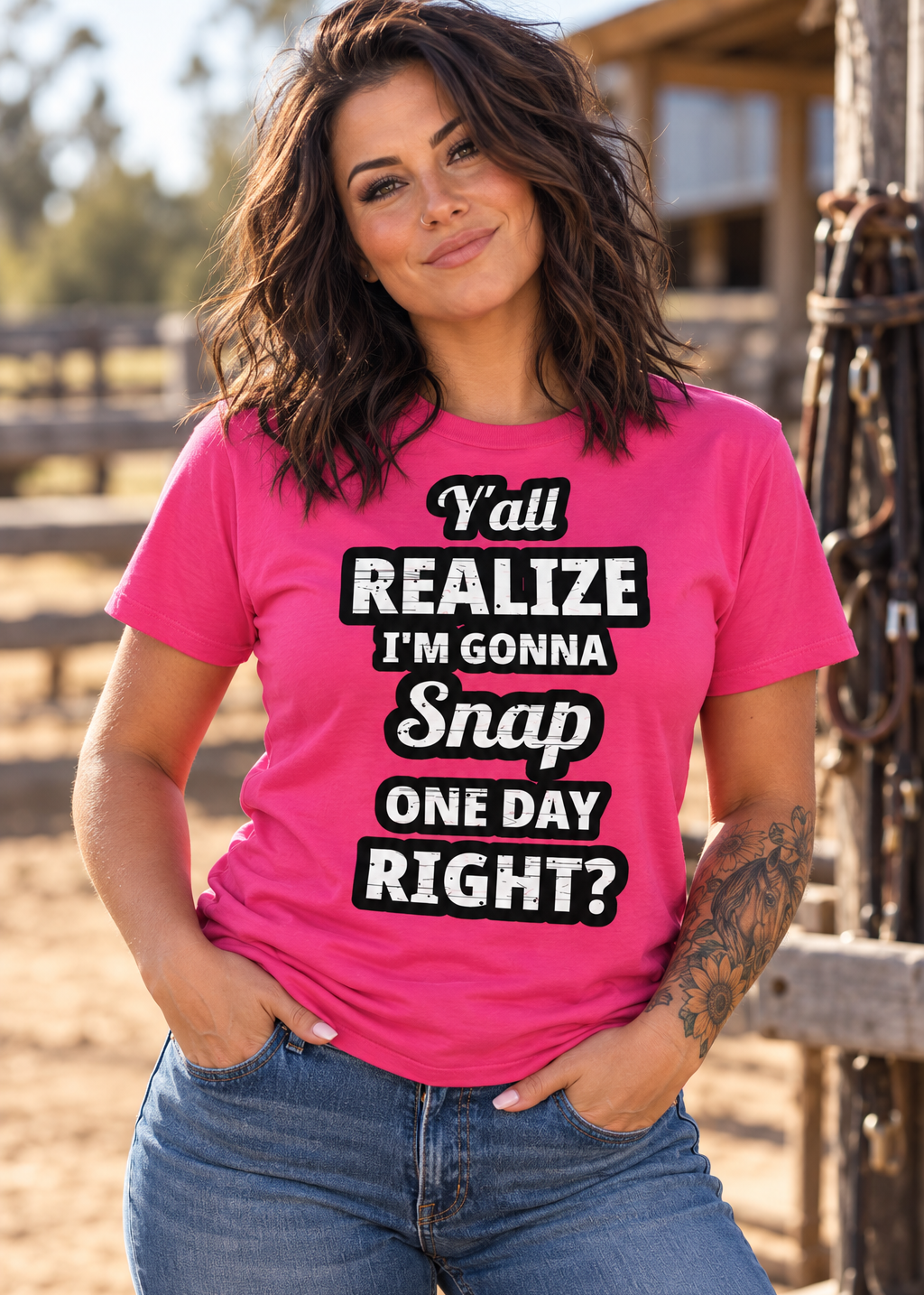 A woman wearing a bright pink graphic t-shirt with the text 'Y'all REALIZE I'M GONNA Snap ONE DAY RIGHT?' in white and black lettering, paired with blue jeans, standing in a rustic outdoor setting.