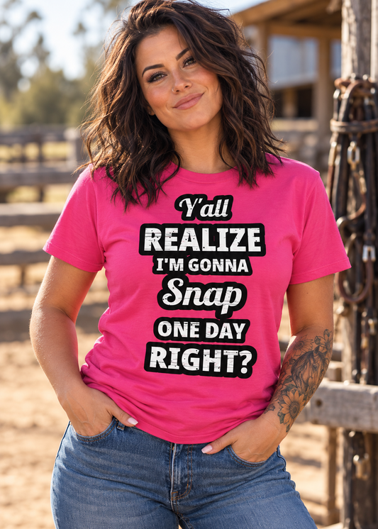 A woman wearing a bright pink graphic t-shirt with the text 'Y'all REALIZE I'M GONNA Snap ONE DAY RIGHT?' in white and black lettering, paired with blue jeans, standing in a rustic outdoor setting.